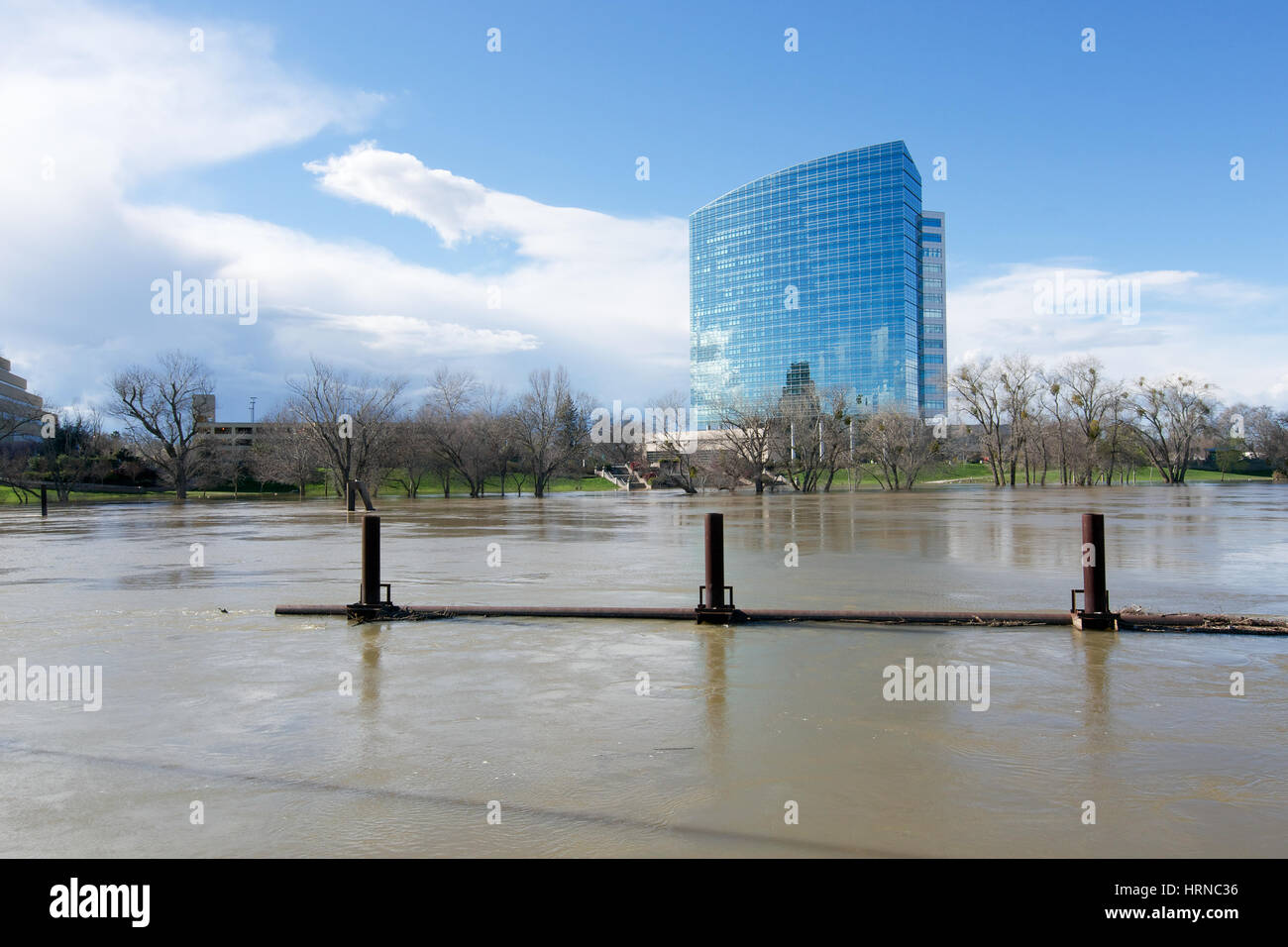 The mirror tower building in West Sacramento viewed from Old Sacramento Historic park on a partly cloudy day, featuring a full Sacramento River inbetw Stock Photo