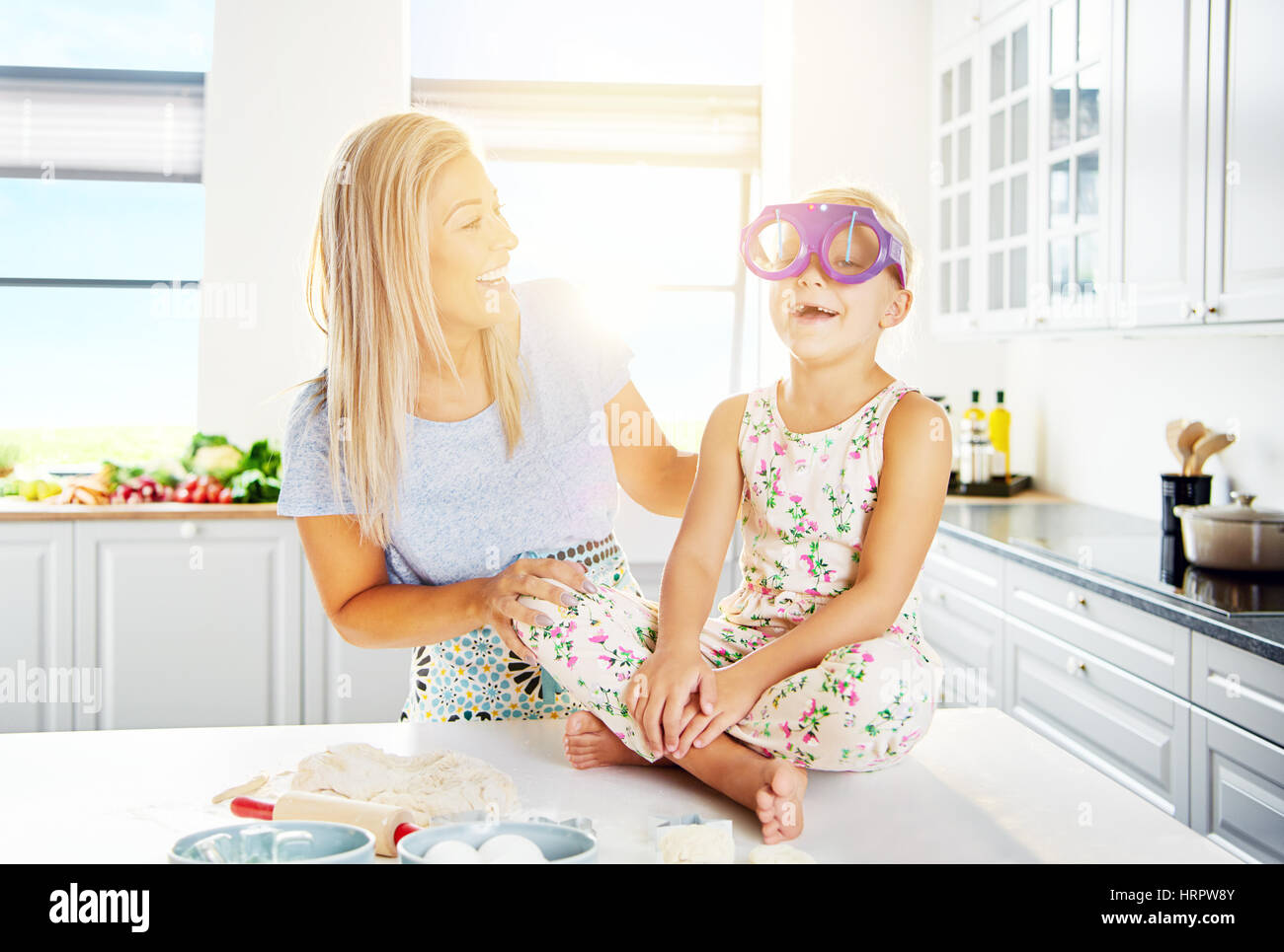 Woman laughing as child wears silly glasses while seated on table beside bread dough ingredients Stock Photo