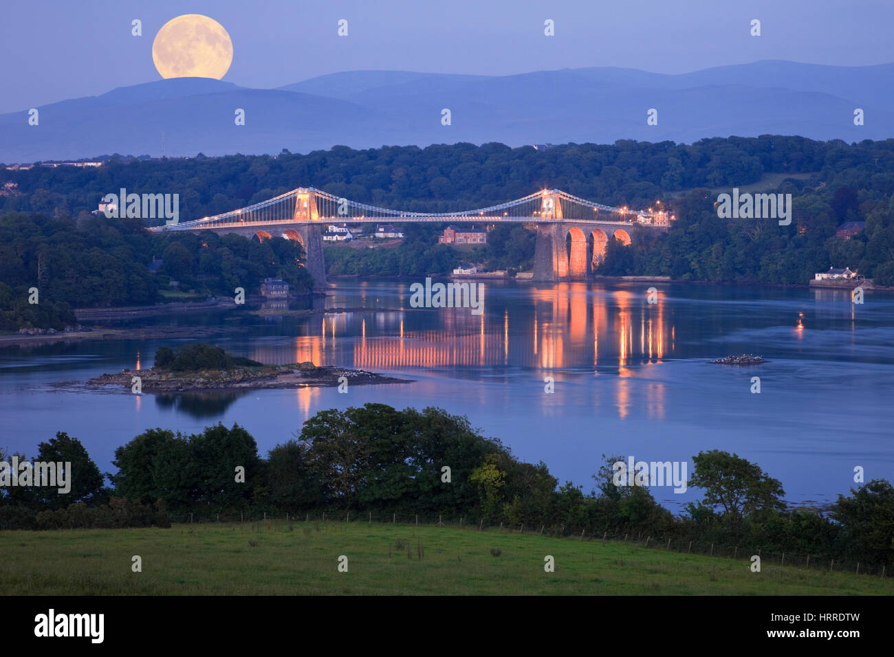 Moonrise full moon rising over Menai Strait coast with Menai suspension bridge illuminated at night. Menai Bridge, Isle of Anglesey, North Wales, UK Stock Photo