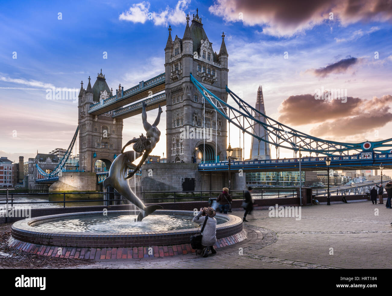 A photographer takes a photograph of  'The  'Girl with a Dolphin' fountain as the sun sets behind Tower Bridge on a calm but cold day in the capital c Stock Photo