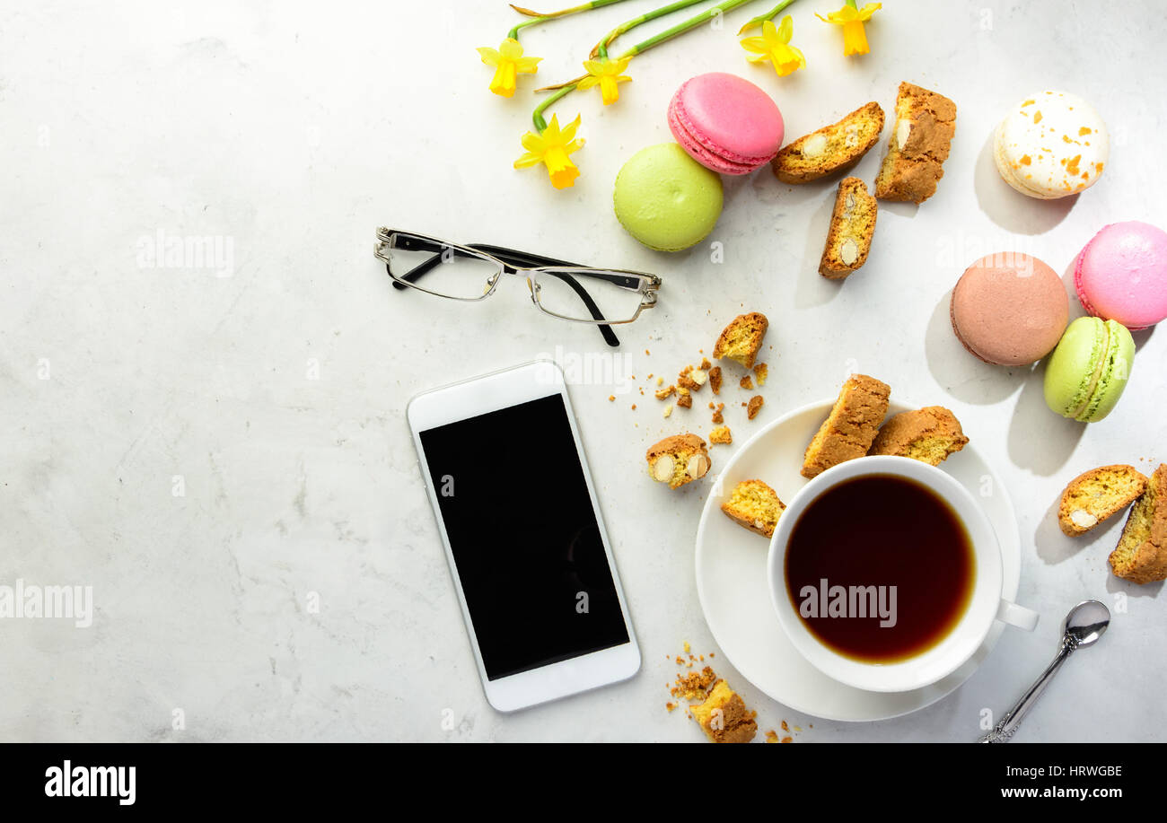 Cup of black tea with tasty almond cookies, rich in vitamins, minerals, varicolored macarons, smartphone, glasses and flowers on a white background in Stock Photo