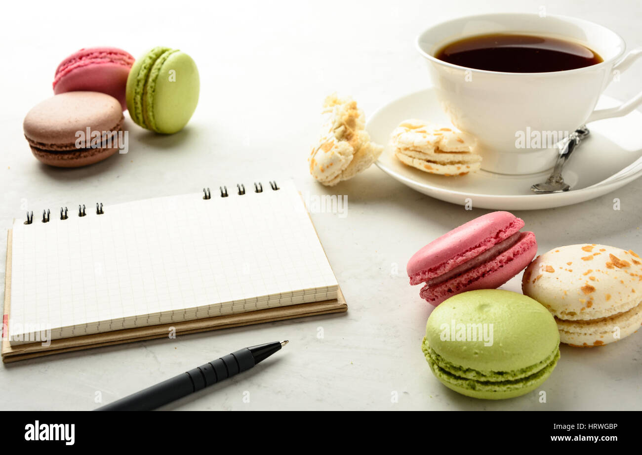 Cup of black tea with tasty varicolored macarons and notebook on a white background in light key. Copy space. Stock Photo