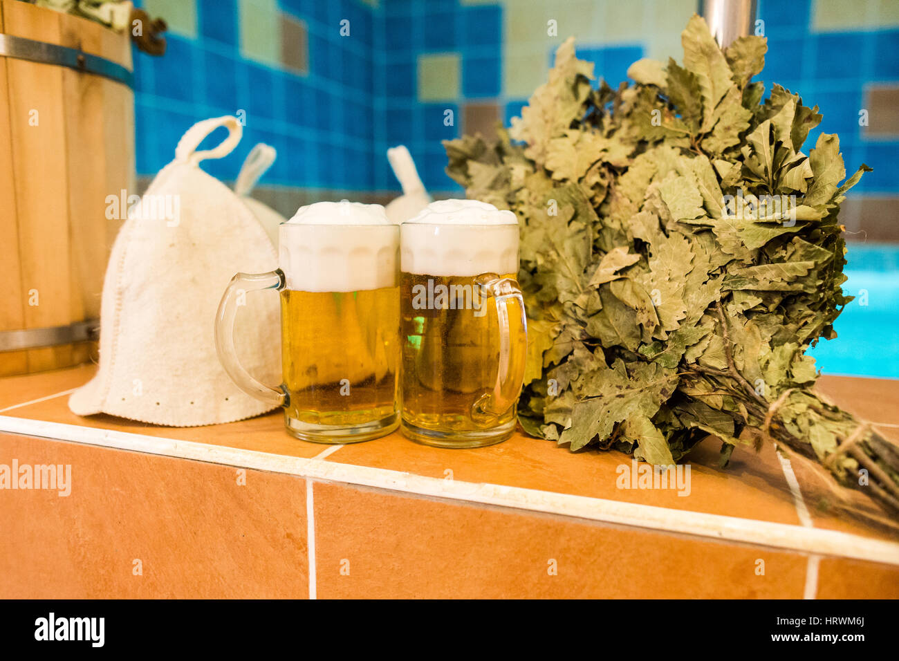 Bath accessories in the Russian bath. Bathroom items of traditional Russian sauna. two mugs of light beer, bathing caps, bath brooms from oak leaves a Stock Photo