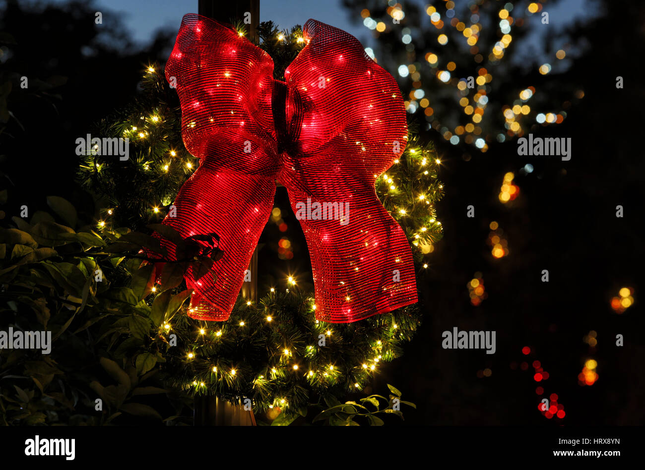 A lighted evergreen Christmas wreath, with a large red bow, displayed on a dark background with christmas lights in the background. Stock Photo