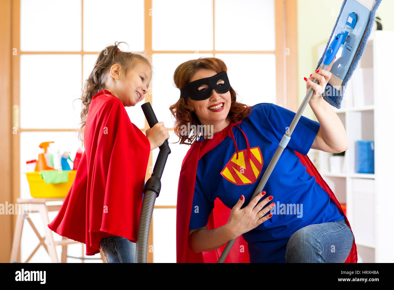 Preschooler girl and her mother dressed like superheroes. Middle-aged woman and kid playing while doing cleanup at home Stock Photo