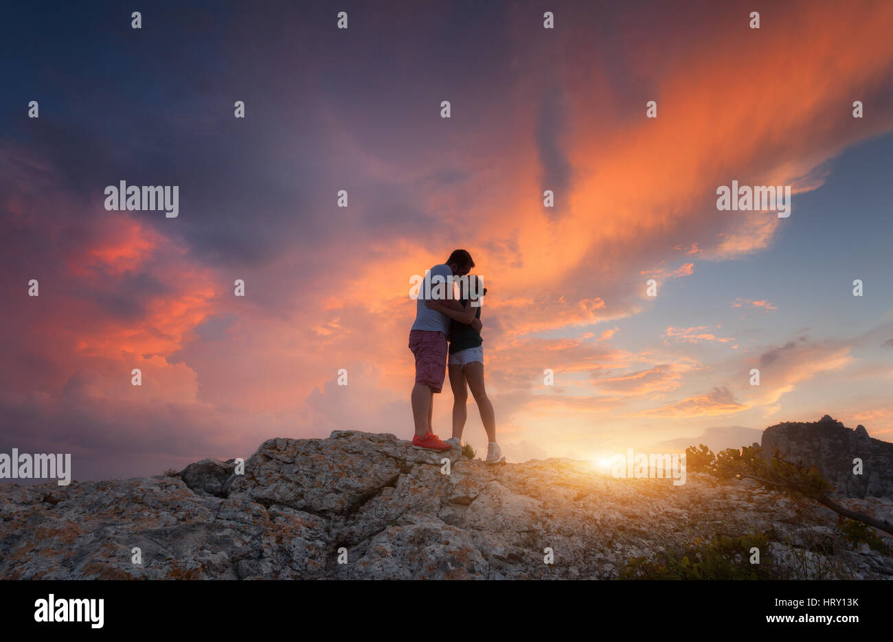 Silhouettes of a hugging and kissing man and woman on the mountain peak at sunset. Landscape with silhouette of young people against colorful sky Stock Photo