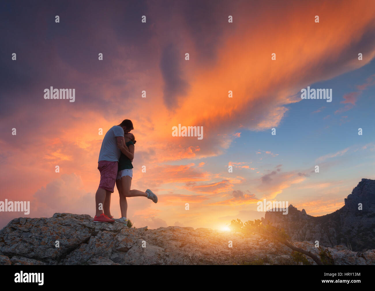 Silhouettes of a hugging and kissing man and girlfriend on the mountain peak at sunset. Man and woman. Landscape with silhouette of young people Stock Photo
