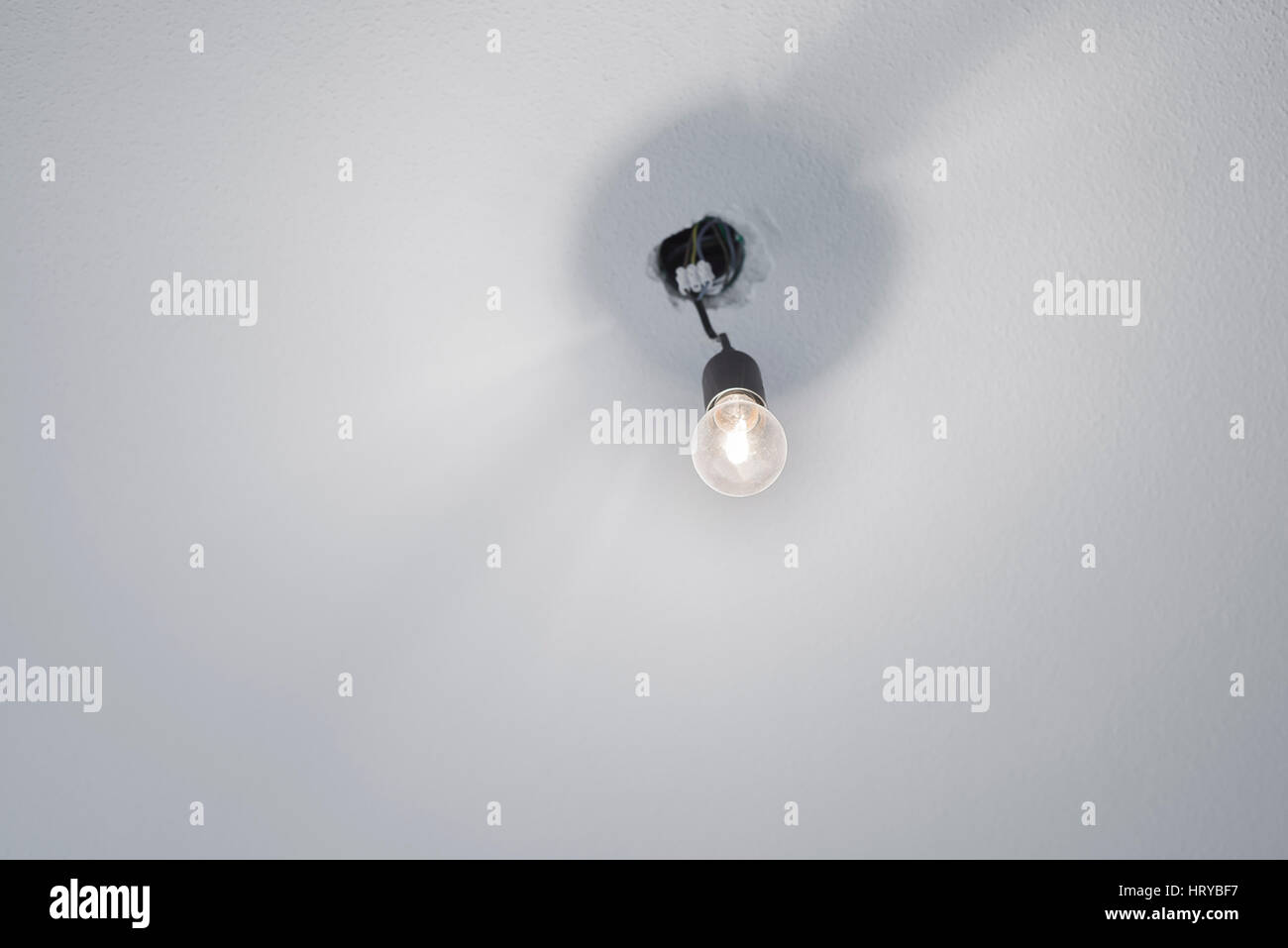 Illuminated halogen incandescent light bulb in a black plastic lamp holder socket with power cables protruding from a ceiling with white rough plaster Stock Photo