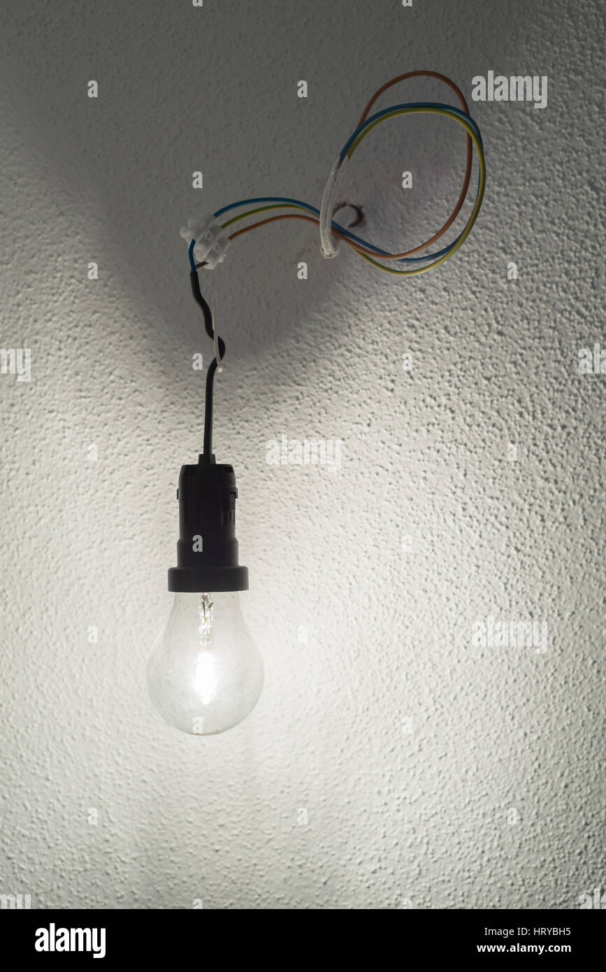 Illuminated halogen incandescent light bulb in a black plastic lamp holder socket with power cables protruding from a wall with white rough plaster Stock Photo