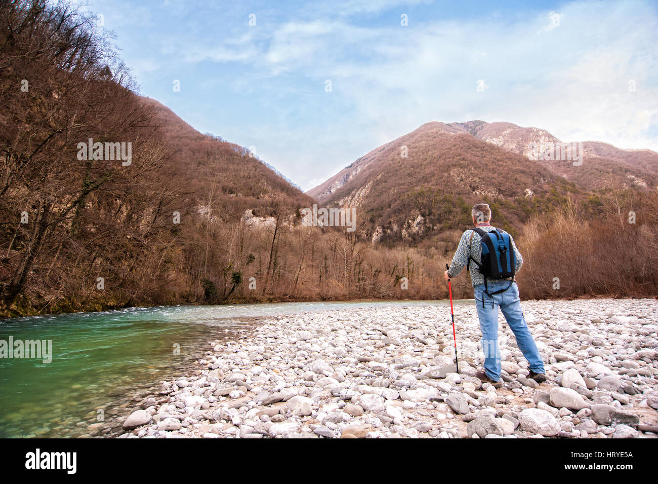 Hiker on the bank of a river. Trekking toward  mountain. Rambler about 60 years old. Stock Photo
