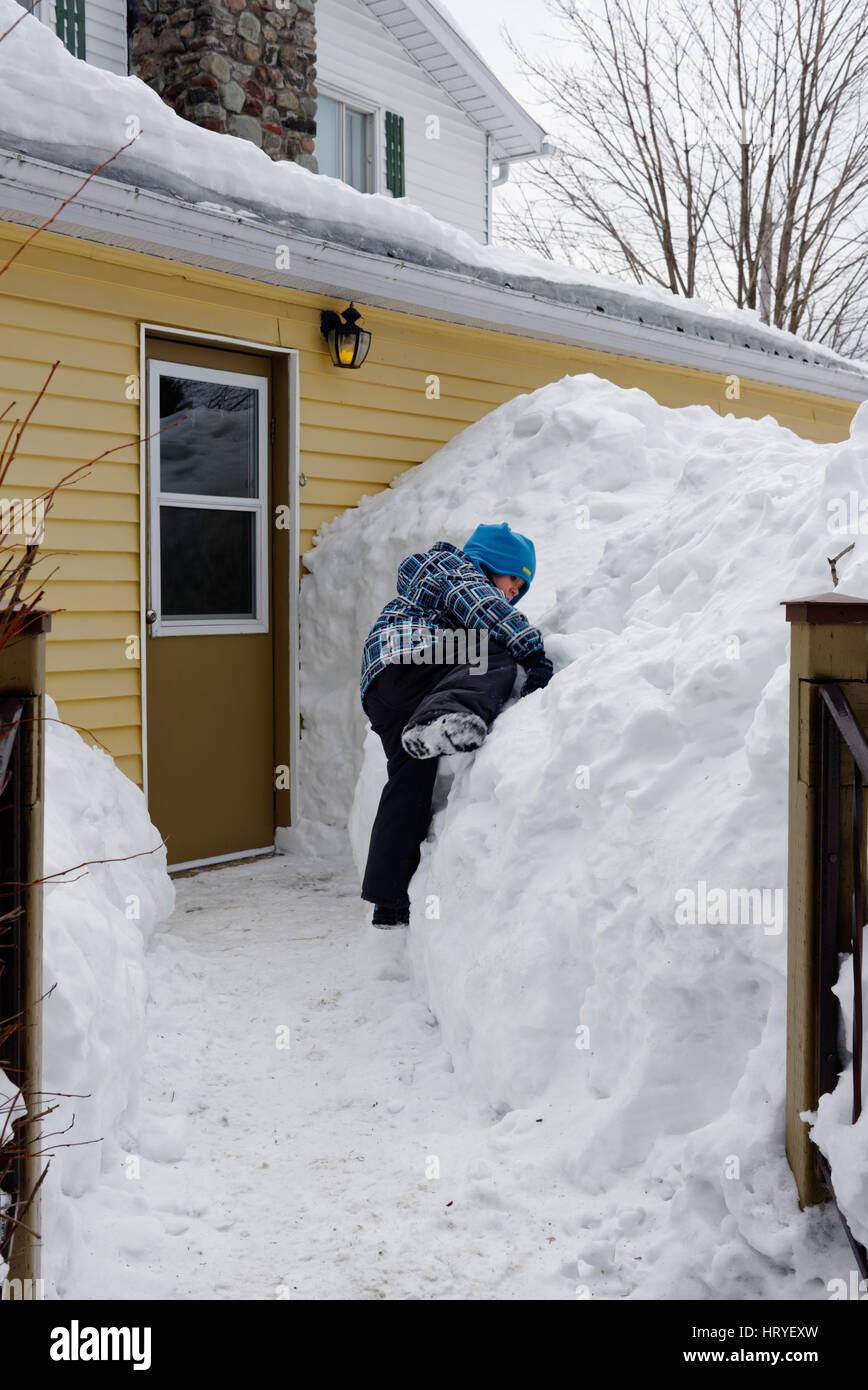 A young boy (4 yr old) climbing on a big snowdrift outside a house in Quebec Stock Photo