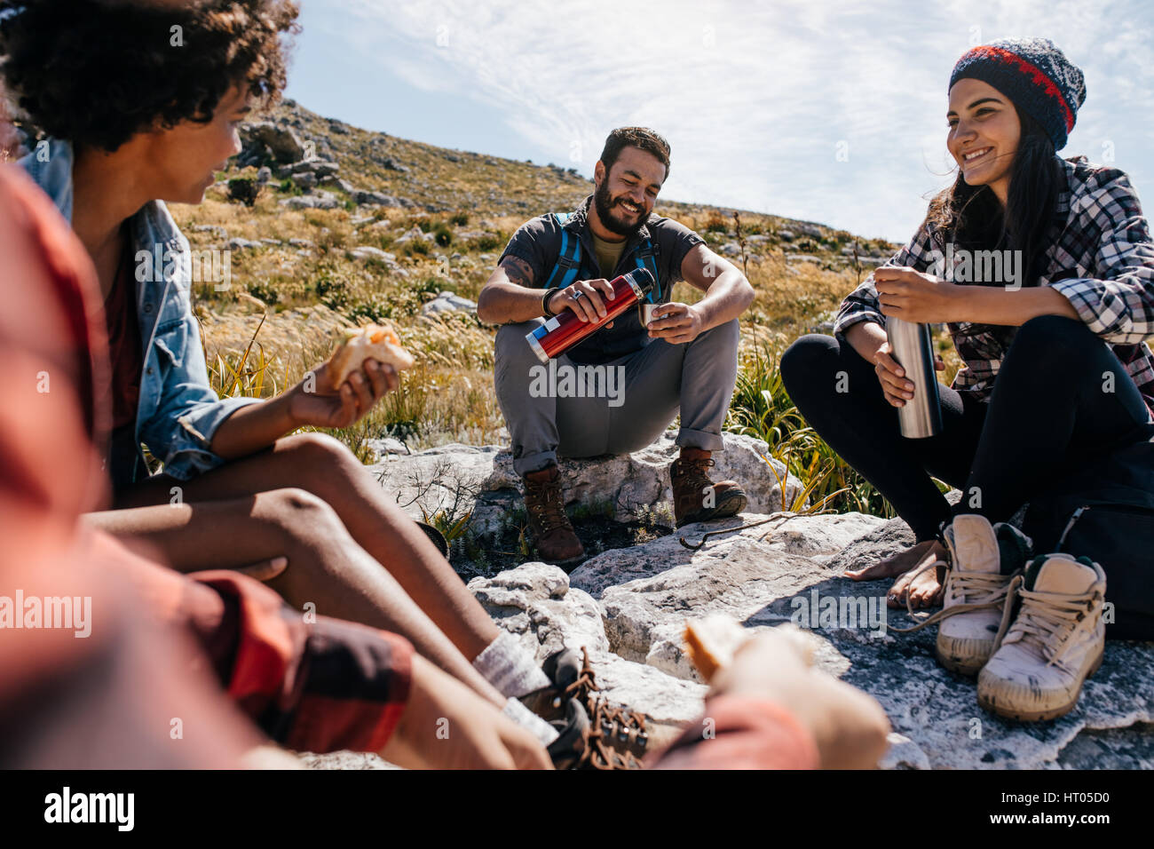 Group of hikers couple eating sandwiches and drinking coffee while relaxing in field. Young people having a rest during country hike. Stock Photo