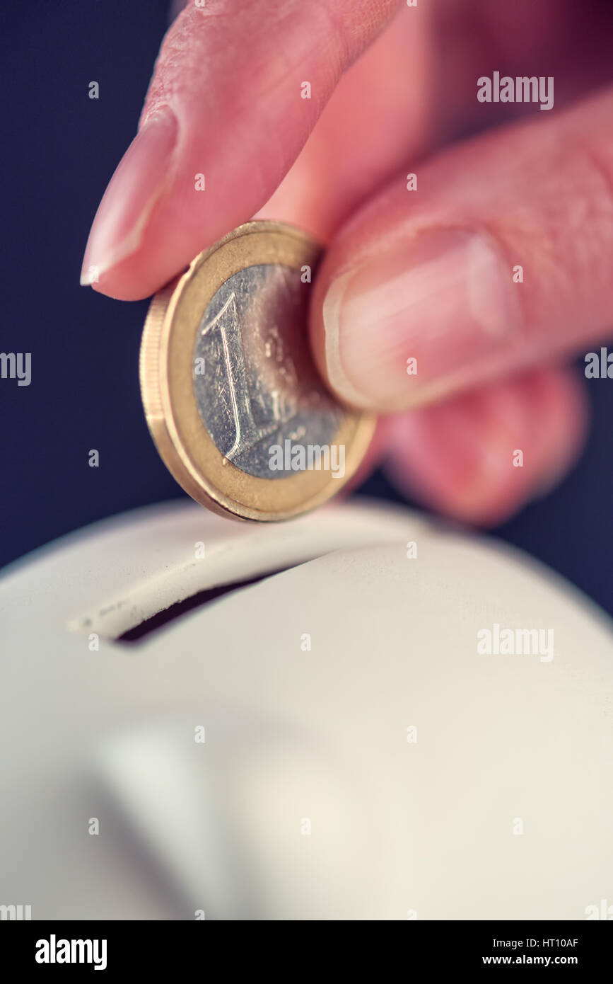 Woman putting one euro coin in piggy bank, money savings and home budget concept, macro close up of fingers Stock Photo