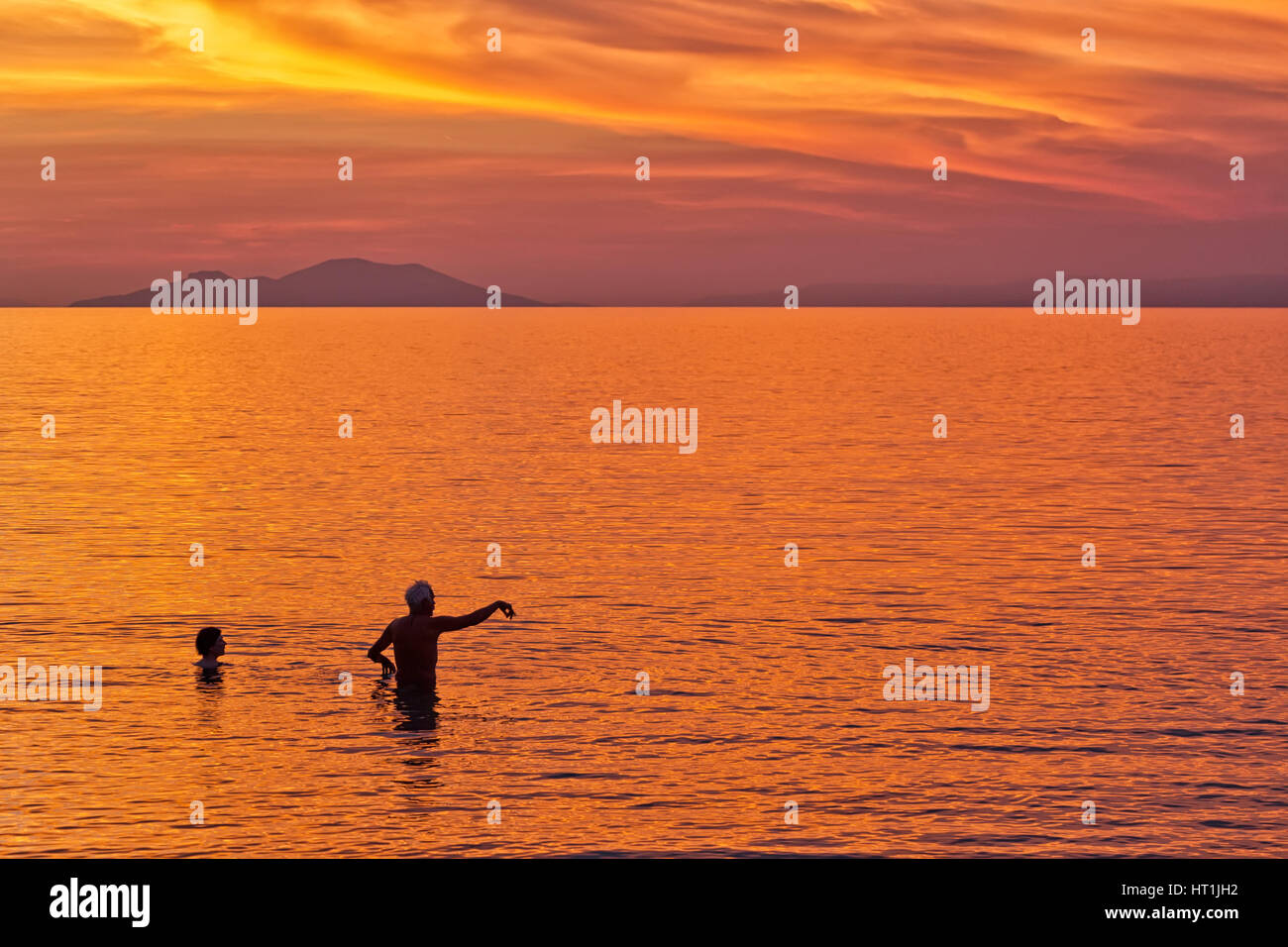 Silhouette of a Couple swimming in the sea against the backdrop of the sun setting on a beach with beautiful dramatic orange and red sky. Stock Photo