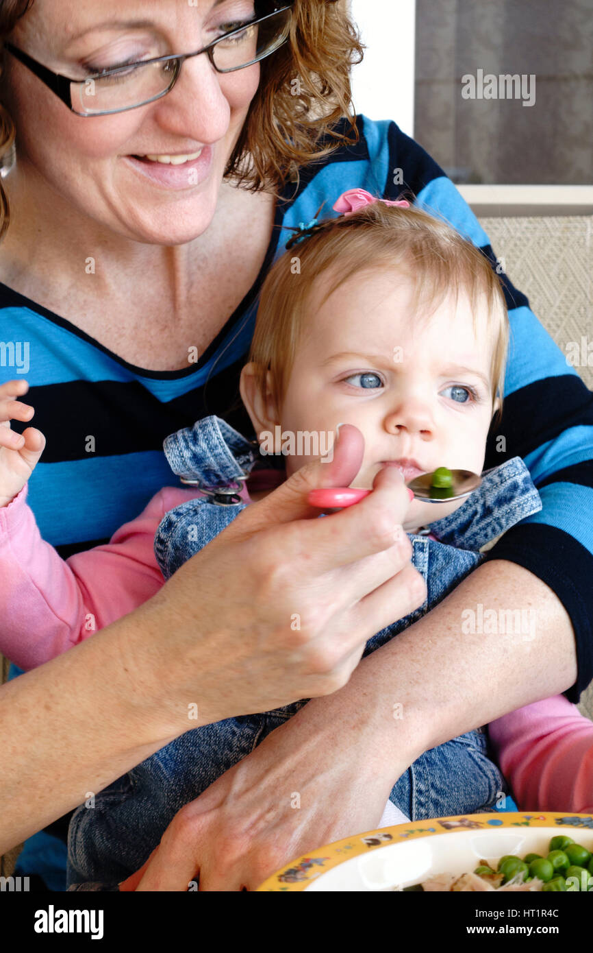 A Caucasian woman grandmother feeds her baby one year old girl grandaughter food from a plate. Kansas, USA. Stock Photo