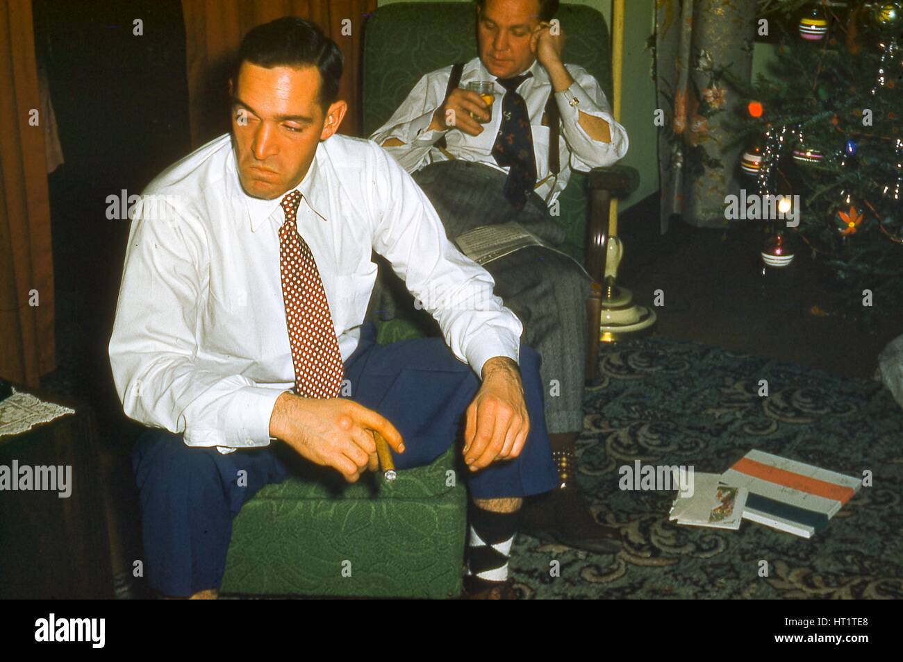 At Christmas, two men sit in a suburban living room near a Christmas tree, one drinking a cocktail, the other wearing a suit, smoking a cigar, and appearing bored, 1955. Stock Photo