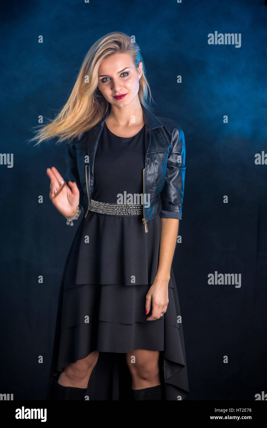 Portrait of attractive blonde woman in black skirt and leather jacket looking at camera in studio shot, on dark background Stock Photo