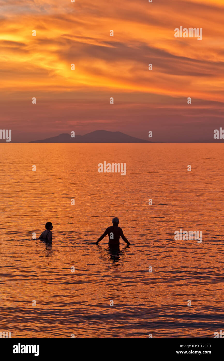 Silhouette of a Couple swimming in the sea against the backdrop of the sun setting on a beach with beautiful dramatic orange and red sky. Stock Photo