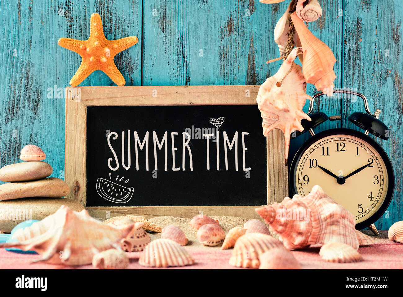 closeup of a chalkboard with the text summer time, an alarm clock, many conches and starfishes, and a beach pail, on a pile of sand, against a bright  Stock Photo