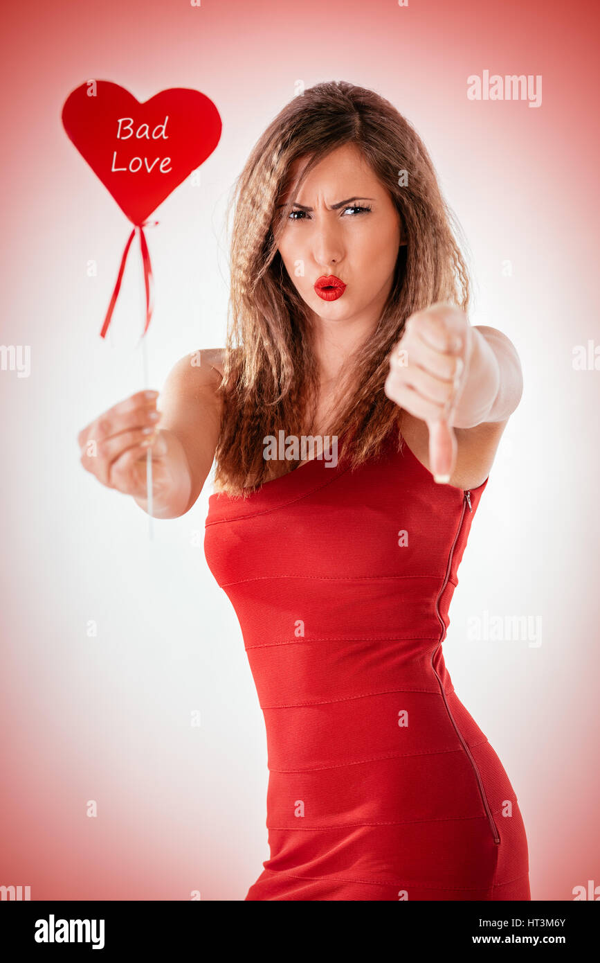 Beautiful displeased girl holding red heart and pointing thumb down. Looking at camera. Valentine's Day concept. Stock Photo