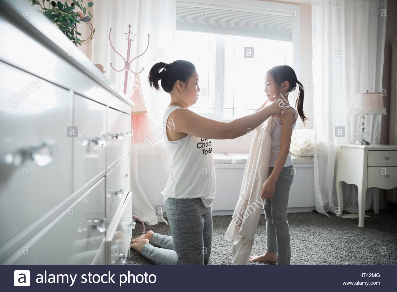 Mother Holding Dress Up To Daughter In Bedroom Stock Photo