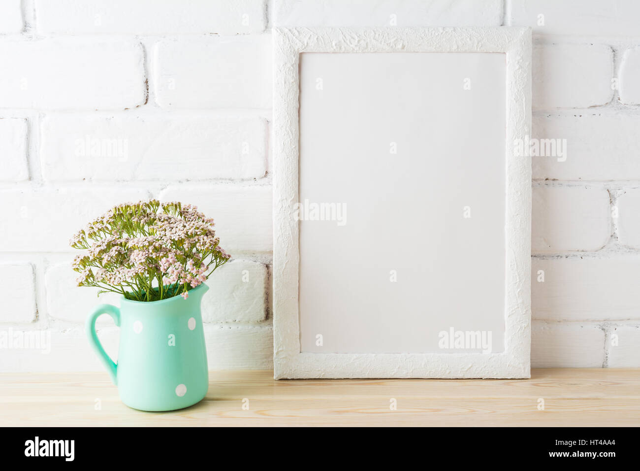 White frame mockup with wild very soft pink flowers in mint pitcher near painted brick wall. Empty frame mock up for presentation design.  Template fr Stock Photo