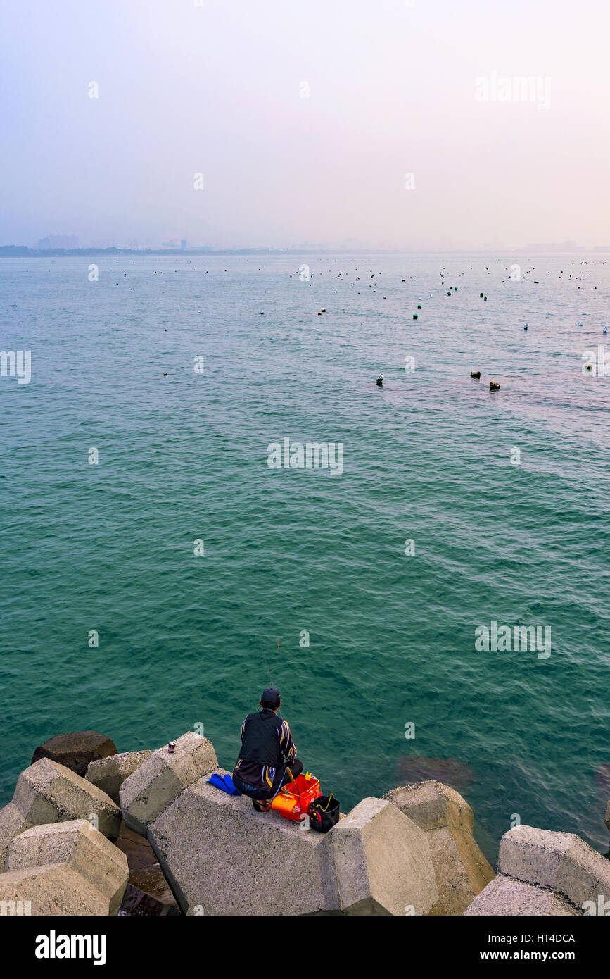 TAIPEI, TAIWAN - JANUARY 05: Fisherman sitting alone fishing on rocks in the Tamsui area of Taipei on January 05, 2017 in Taipei Stock Photo