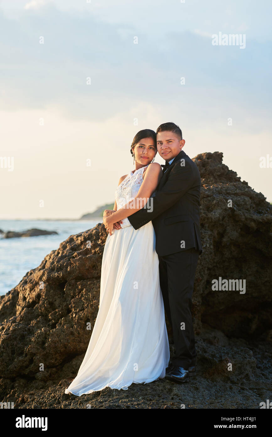 Hispanic bride and groom standing on rock coastline at sunset orange light. Latino couple in love wedding day Stock Photo