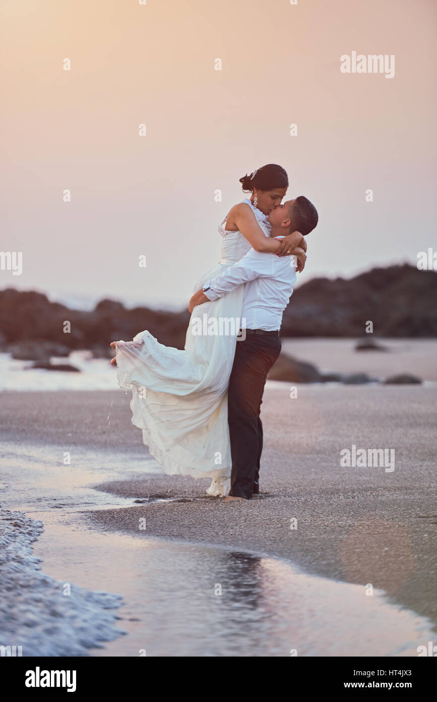 Happy kissing groom and bride stand on ocean coastline in sunset light ...