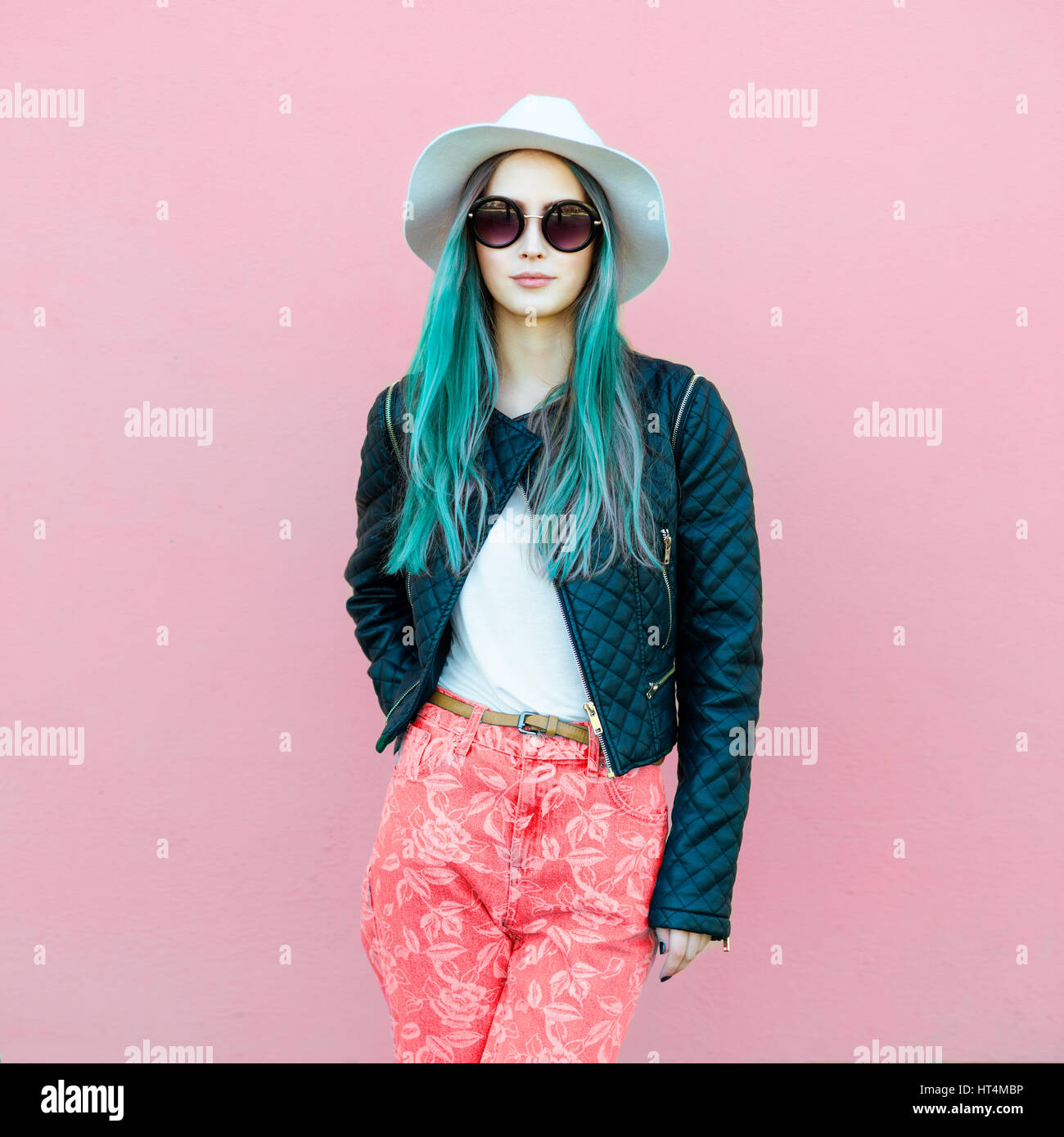 Fashionable young blogger woman with blue hair wearing casual style outfit with black jacket, white hat, pink jeans and sunglasses posing near the wal Stock Photo