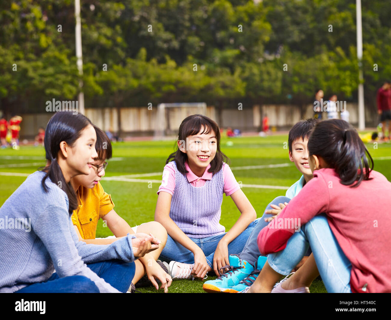 group of asian elementary school boys and girls sitting and chatting on playground grass Stock Photo