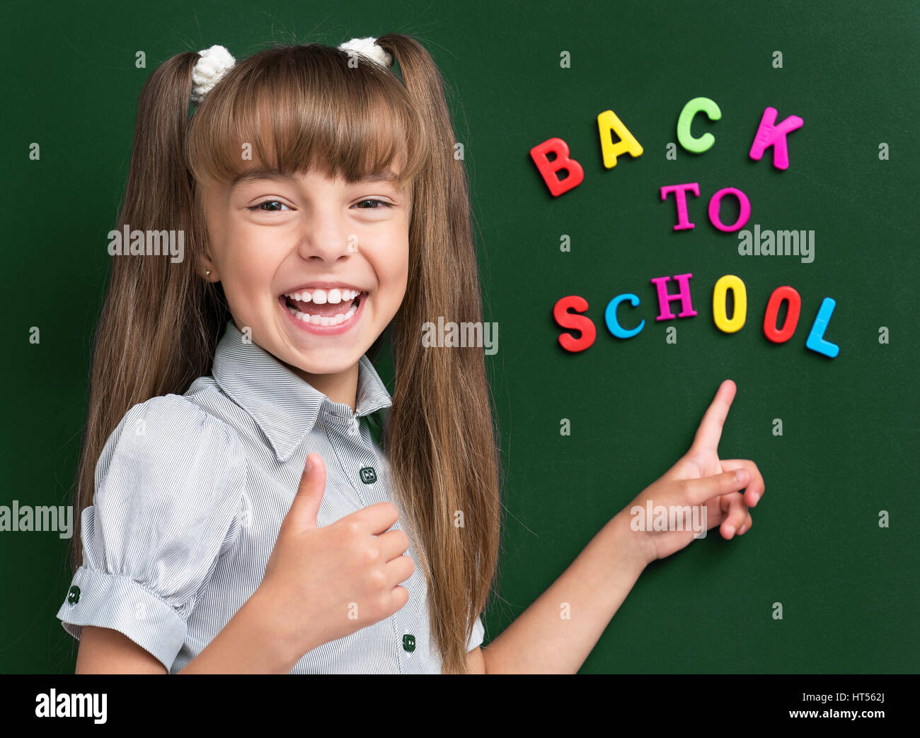 Portrait of adorable young girl showing thumb up sign at the green chalkboard in classroom. Stock Photo