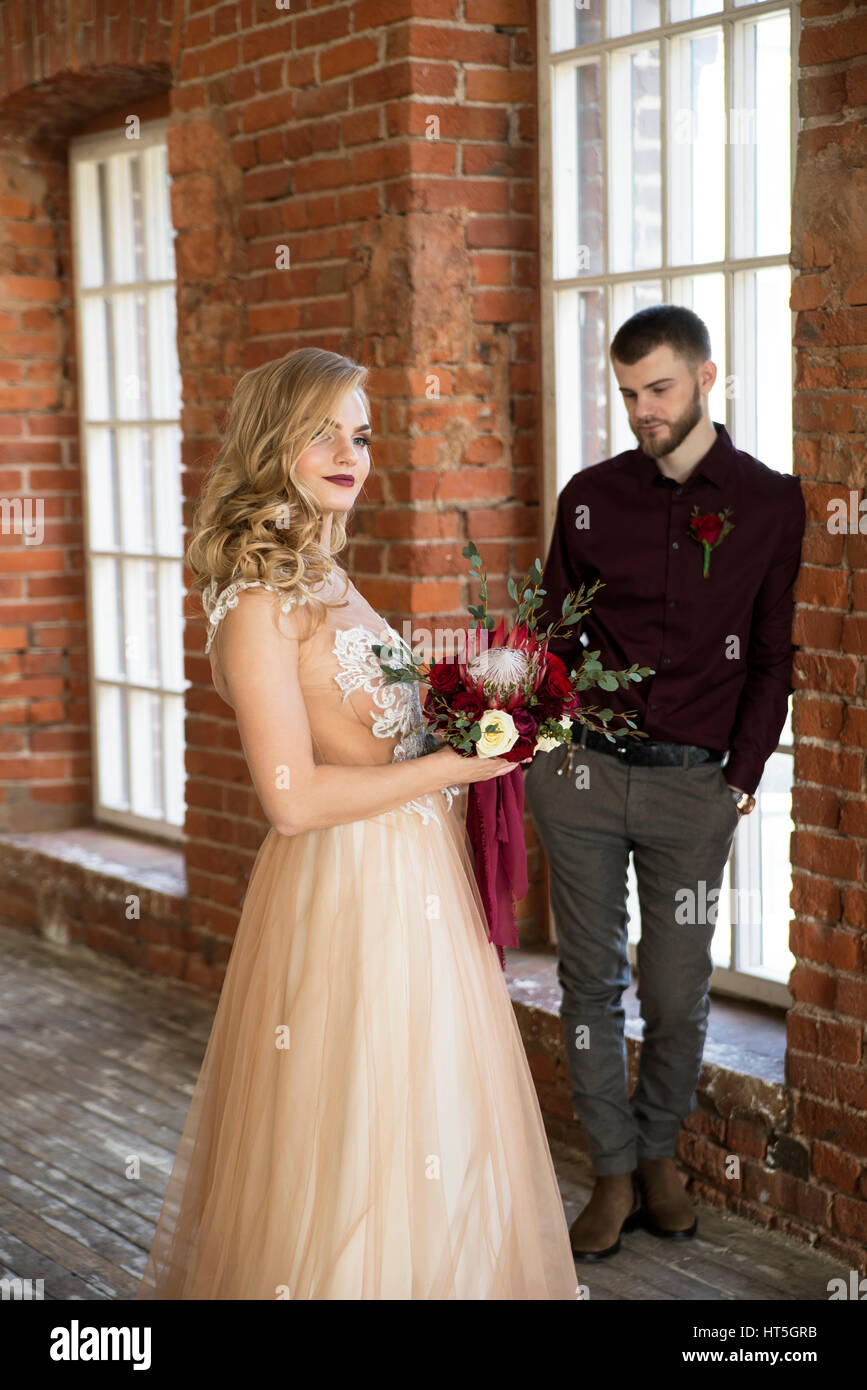 bride in a beautiful beige dress with bouquet and groom in a Burgundy shirt with a boutonniere pose in front of the window at a brick wall Stock Photo