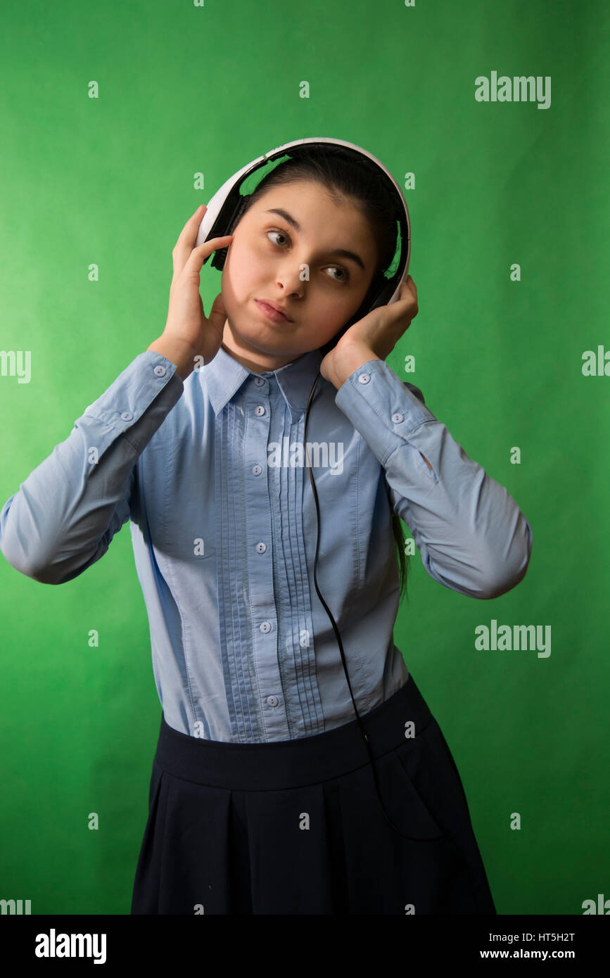 teen schoolgirl in blue shirt is listening to music in white headphones against a green background Stock Photo