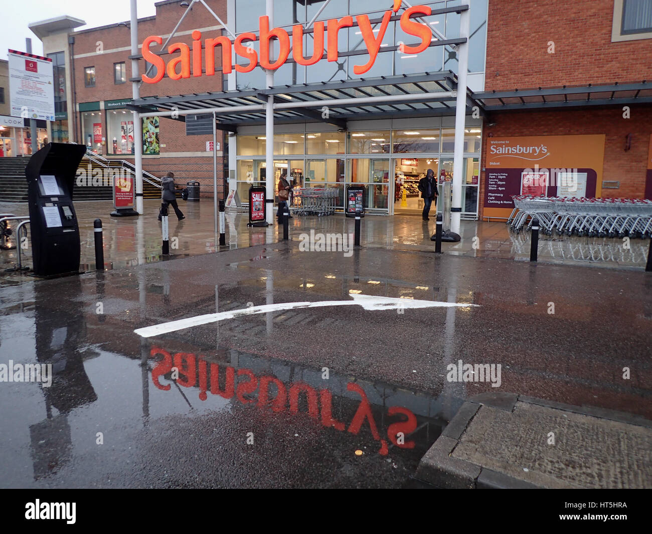 The entrance of Didcot Sainsbury's in rain with the Sainsbury's sign reflected in a puddle in front of the entrance in full Stock Photo