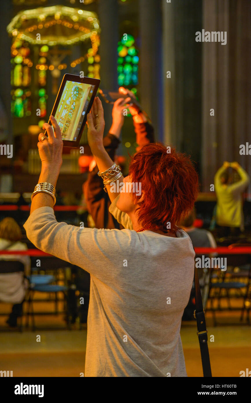 A portrait format image of a red haired woman taking a photo with her ipad in the Sagrada Familia church in Barcelona, Spain. Stock Photo