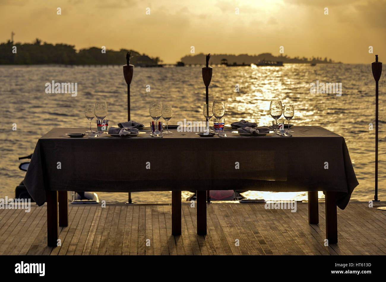 A dining table set against the setting sun, near an island, seen from the deck of a ship. Stock Photo