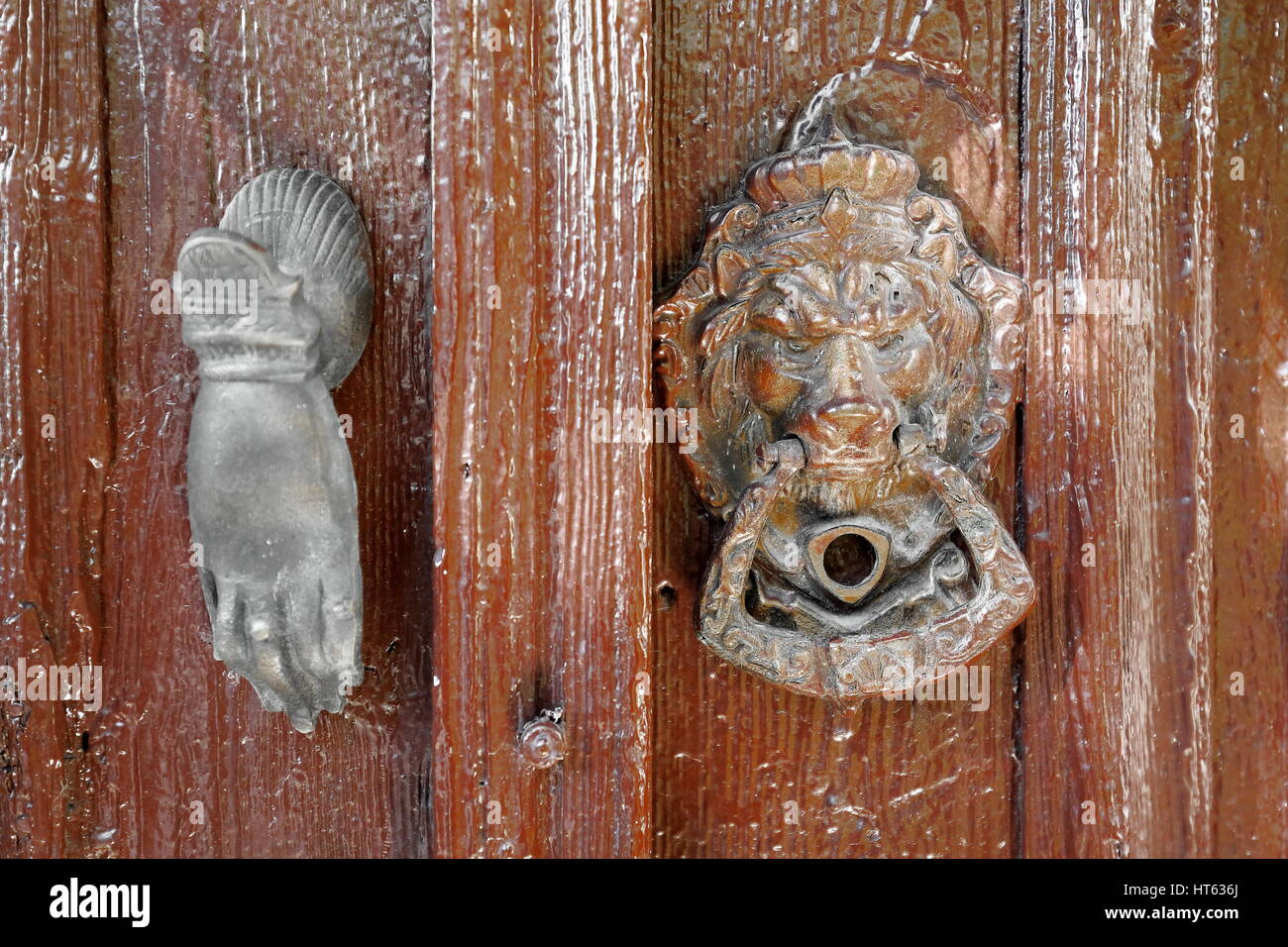 Old metallic doorknockers in the shape of a lionhead and a human hand placed on a brown painted-old woooden door of a house in the central area of tow Stock Photo