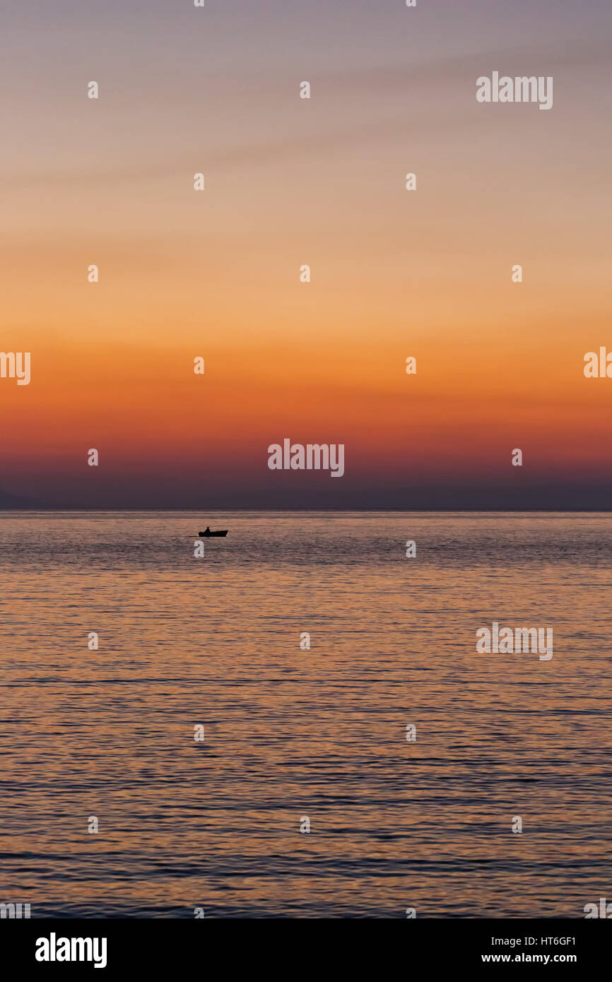 Small boat sunset silhouette is a fisherman's boat paddling along the water with a colorful purple and pink night sky in the background Stock Photo