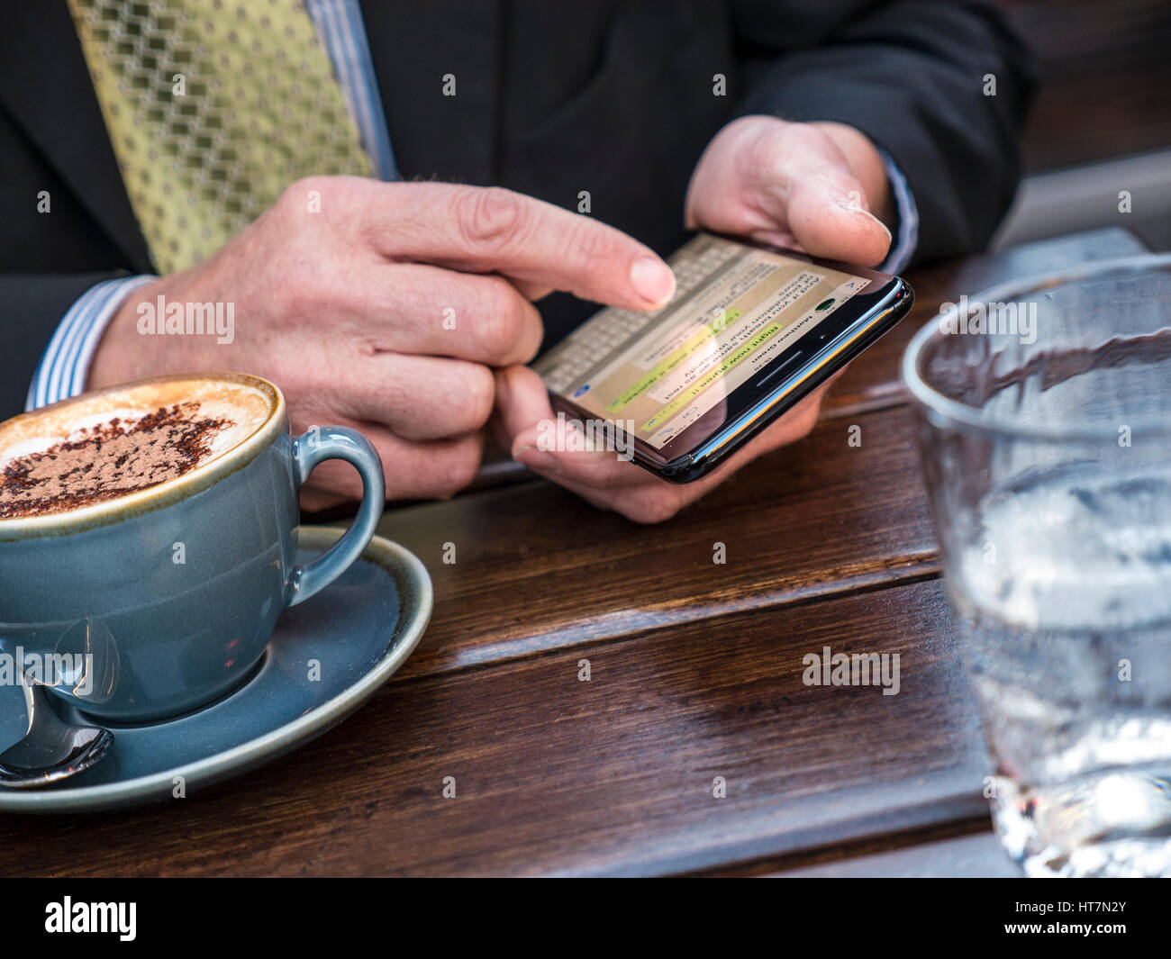Businessman texting mature businessman's hands close texting with smartphone sitting at alfresco restaurant with glass of water and cappuccino coffee Stock Photo