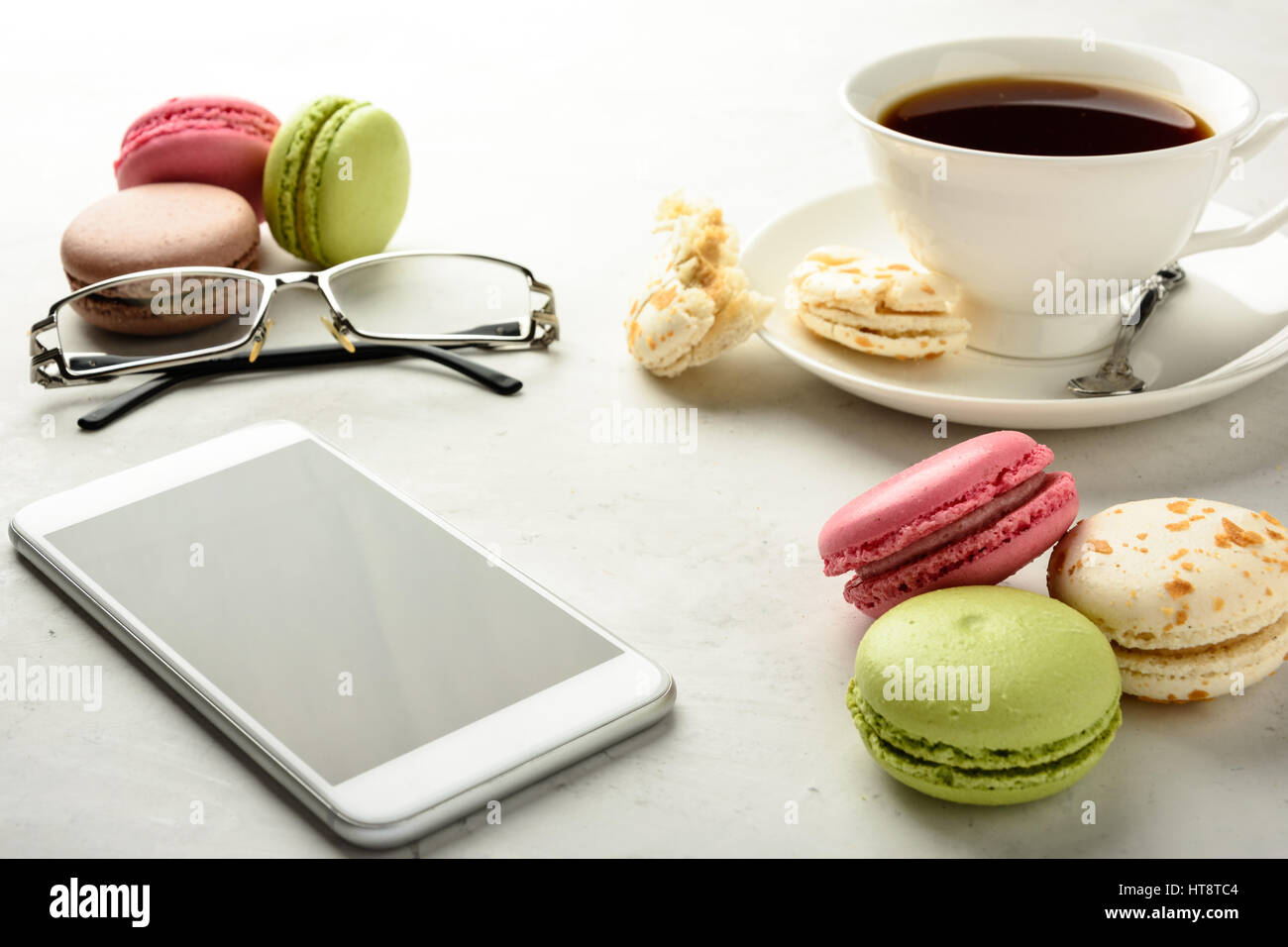 Cup of black tea with tasty varicolored macaroons, smartphone, glasses on a white background in light key. Stock Photo