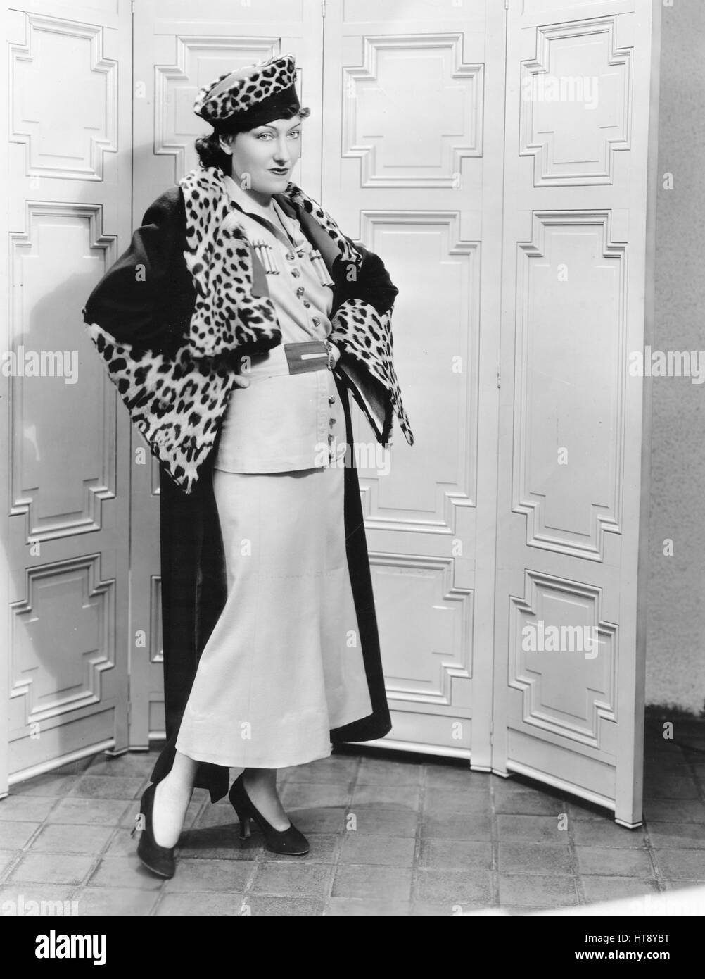 'Strictly Russian is this new ensemble worn by Gloria Swanson, charming Metro-Goldwyn-Mayer star. The dress is of gold woolen trimmed with cartridge pleating and red banding. The sleeves are full, caught in at the wrist by tight cuffs. The coat is of brown woolen lined with the gold. There is a red border around the lining. Leopard skin trims the collar and sleeves. The hat is a cossack type of leopard and matching material.' Stock Photo