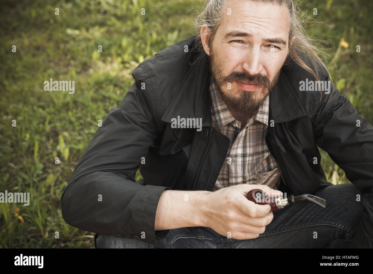 Young asian bearded man smoking pipe in summer park, closeup portrait Stock Photo