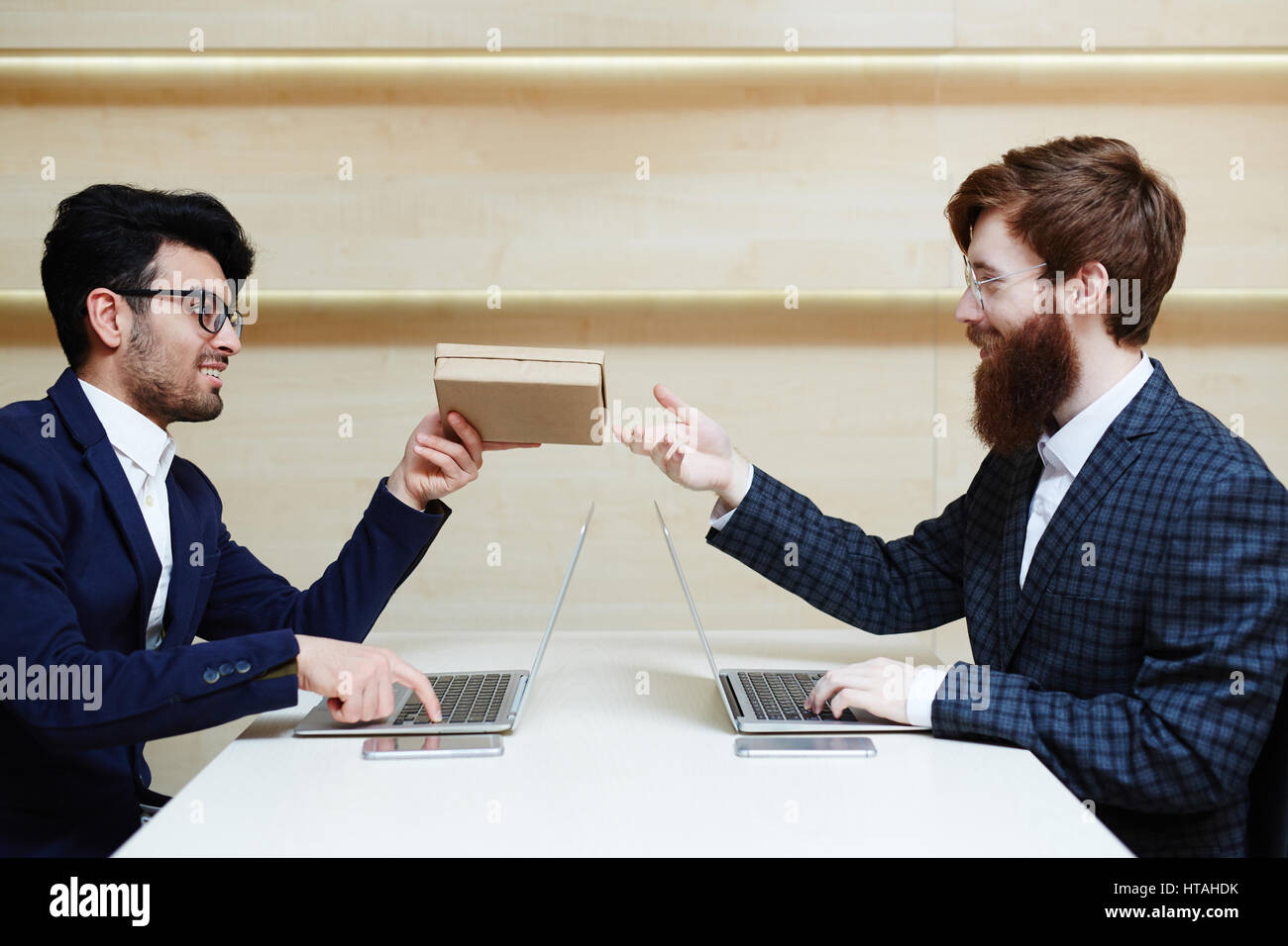 Birthday wishes: two bearded businessmen in eyeglasses working on laptops in office, one of them distracting another for a second in order to give him Stock Photo