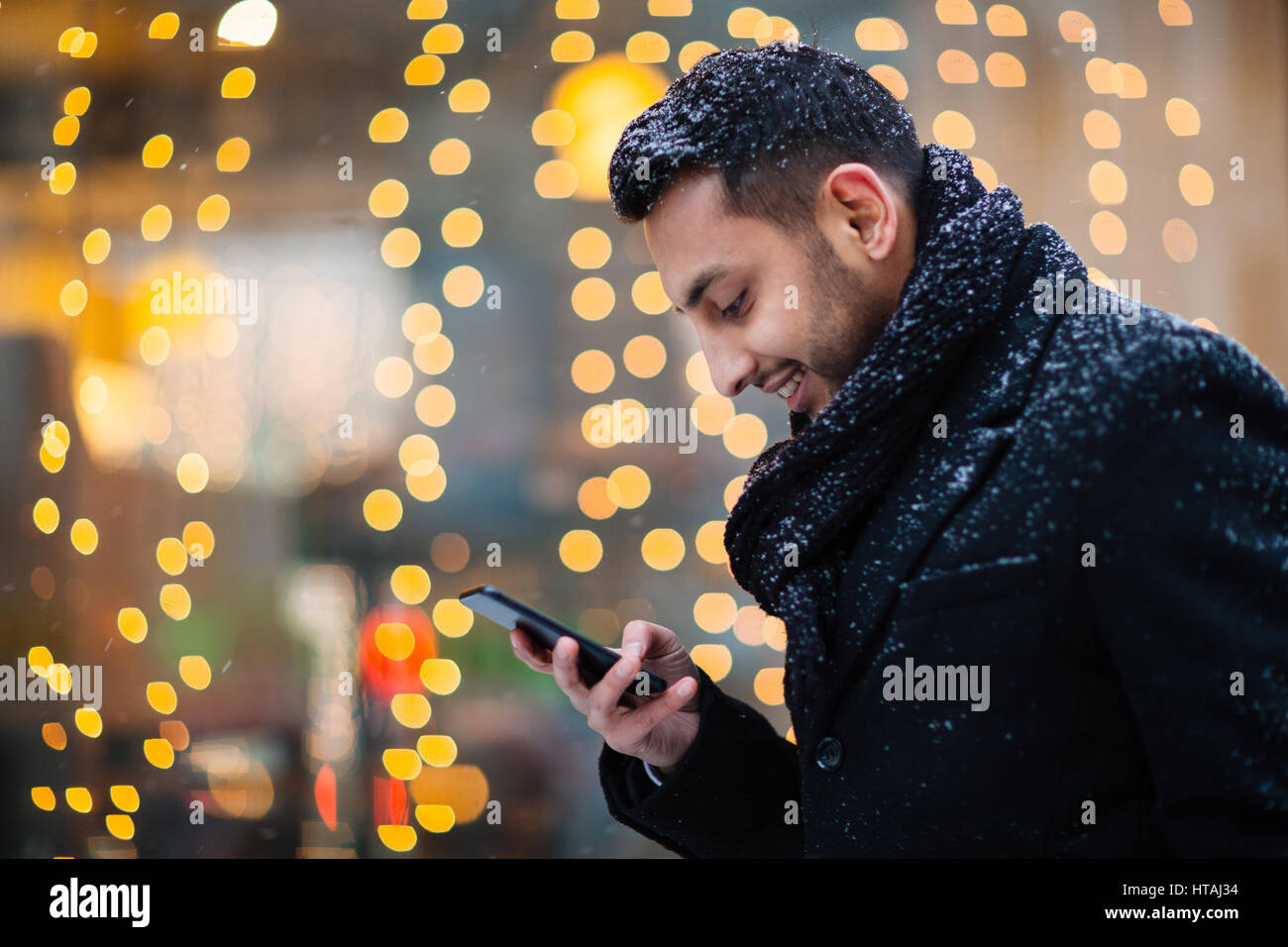 Heartwarming image of young Arabic man stopping to read pleasant text message in the streets of winter city on background of store window decorated by Stock Photo