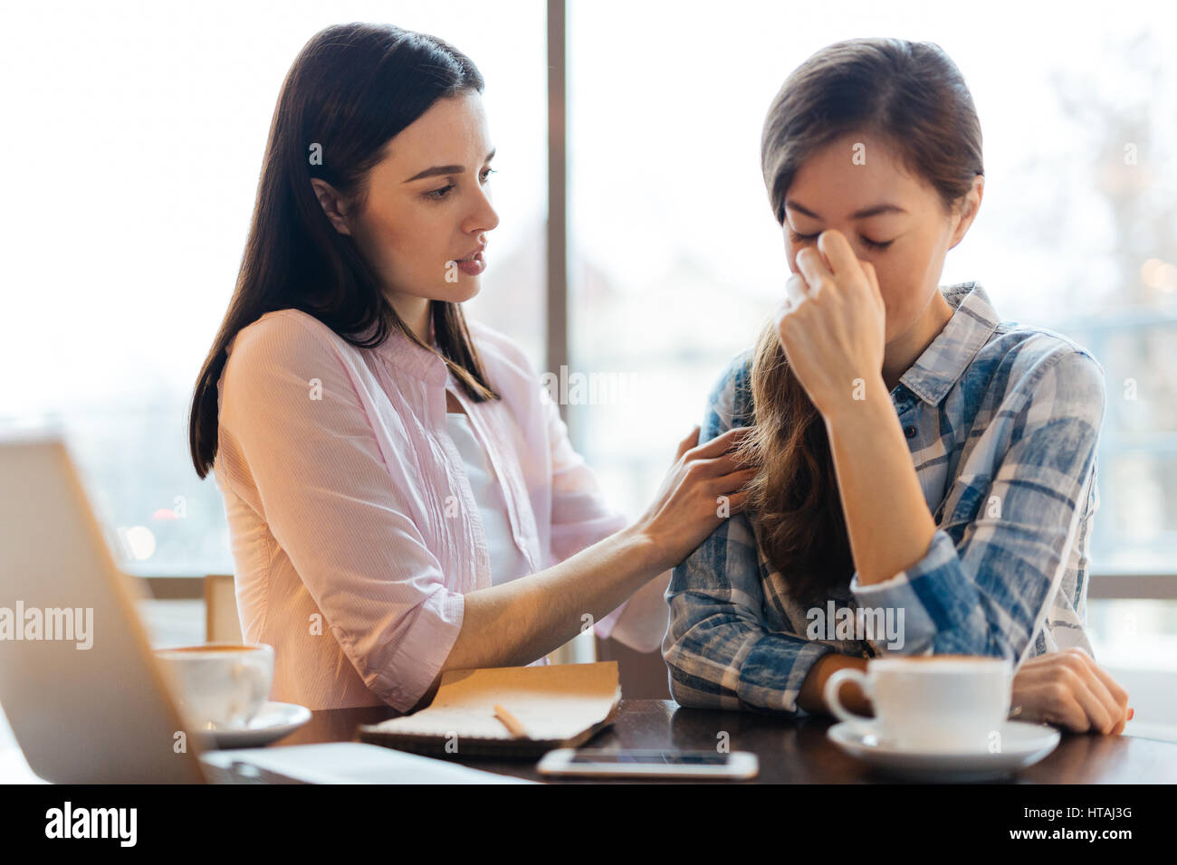 Sad young woman hiding her face crying at table in cafe and her friend hugging her by shoulders in compassion, giving advice trying to console her Stock Photo