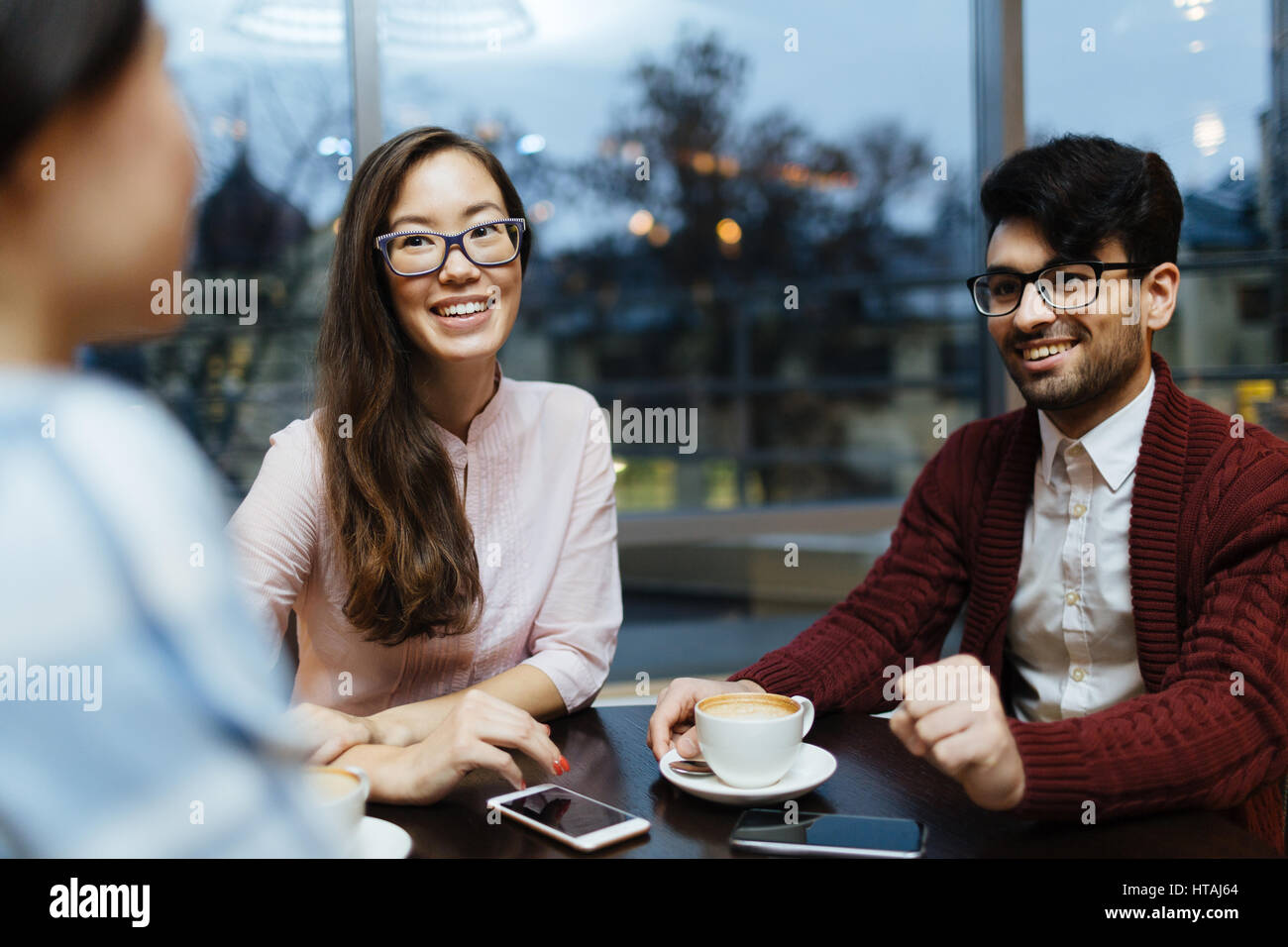International group of young people sitting round table in cafe against window with view to evening city streets, focus on pretty Asian woman and midd Stock Photo