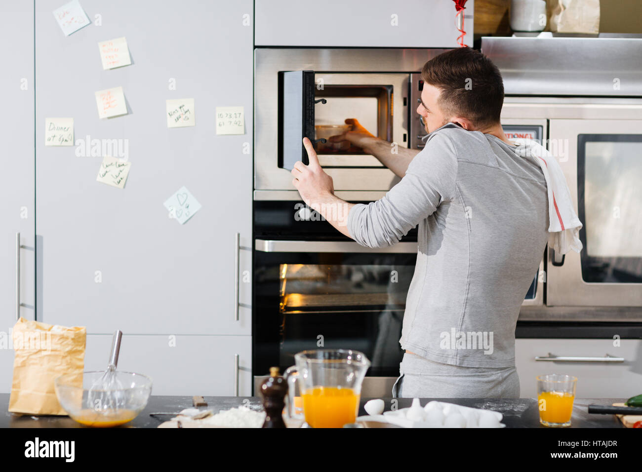 Young man taking out container with hot food from microwave oven while talking on smartphone Stock Photo