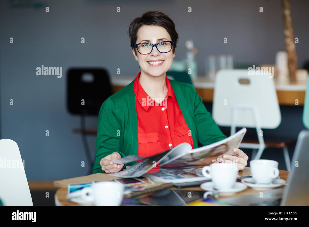 Portrait of young successful businesswoman wearing colorful casual clothes smiling cheerfully at camera while reading magazines at table Stock Photo