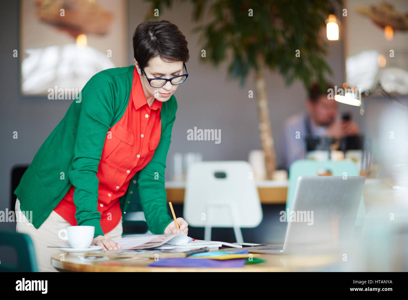 Portrait of young successful businesswoman wearing colorful casual clothes standing at table working with notes and magazines Stock Photo